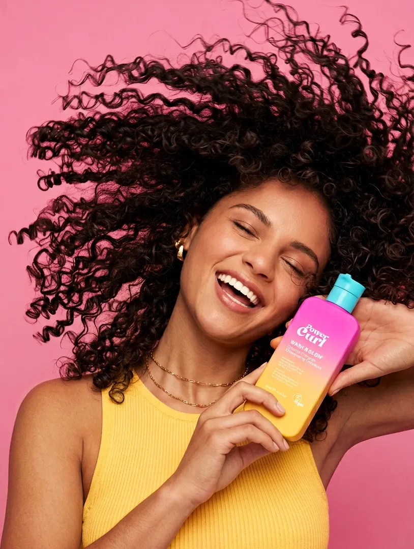 Smiling woman flipping her curly hair while holding Power Curl Wash & Glow shampoo